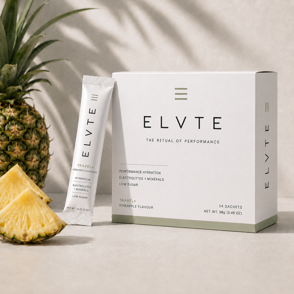 ELVTE Travel+ electrolyte powder sachets shown with a white product box and one standing sachet, styled on a light beige surface with a pineapple slices in the background.