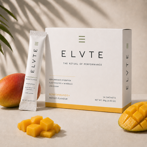 ELVTE Performance+ Hydration sachet and box packaging displayed on a beige surface with mango fruit and leaves around it. The white box and sachet feature black text and a yellow-orange accent band, labeled Mango Flavour.