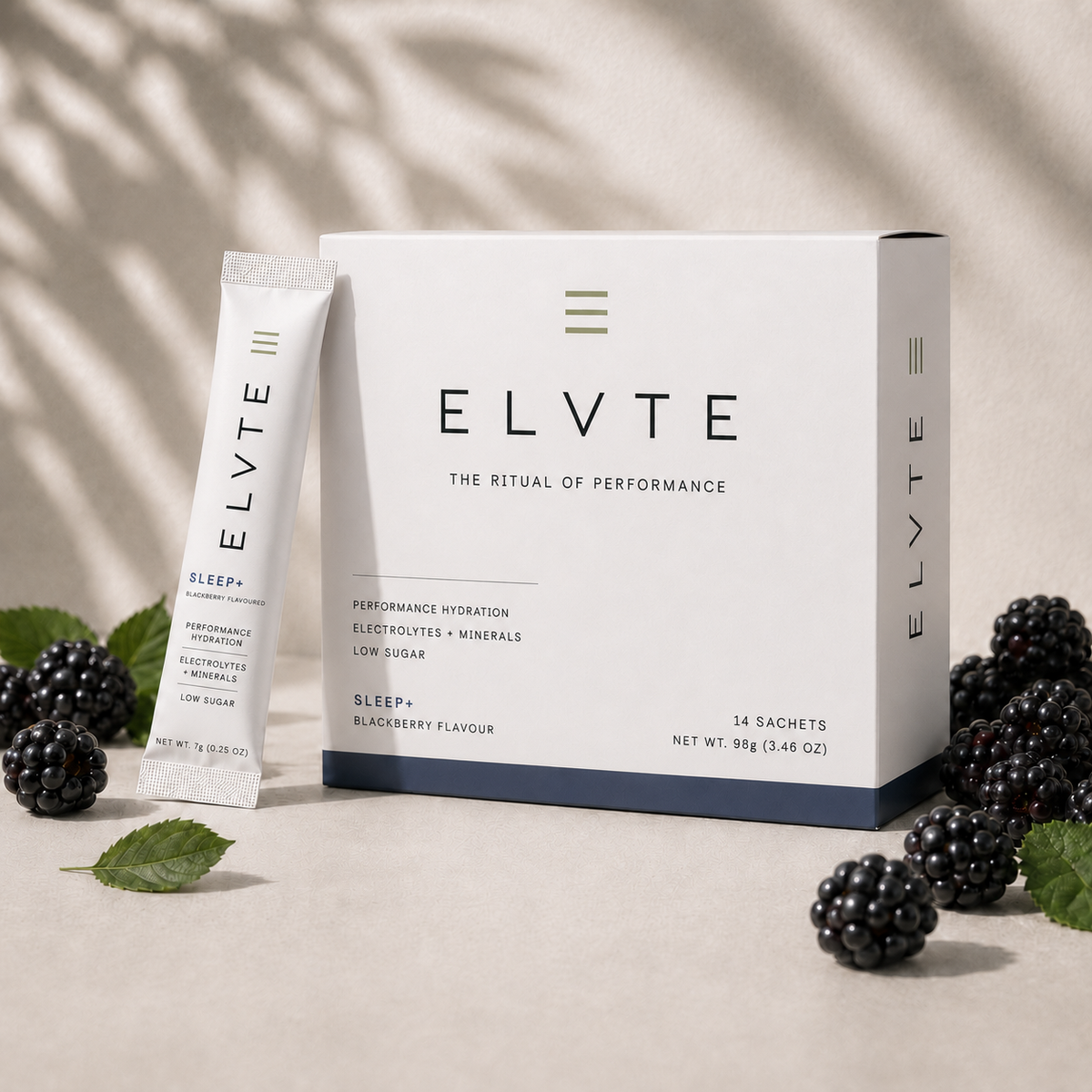ELVTE Sleep+ performance hydration product shown with a white box, one single sachet, and blackberries on a beige surface with soft leaf shadows. The box and sachet are white with black text and a small green accent line.